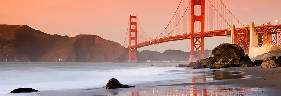 Solnedgång bakom Golden Gate-bron med lugnt vatten och strand i förgrunden.