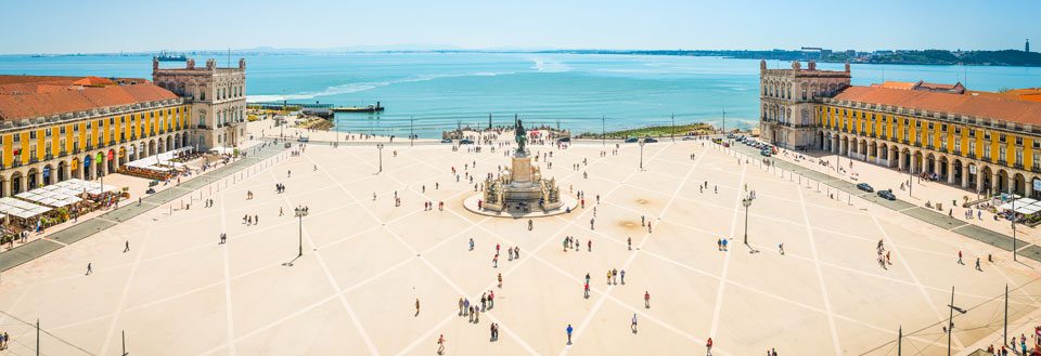 Panoramavy över ett rymligt, öppet torg i Lisboa med promenerande människor, omgiven av klassiska byggnader och havet i bakgrunden.