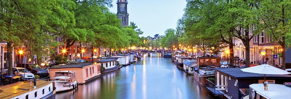 Bilden visar en kanal i Amsterdam i skymningen. Hus båtar är parkerade längs kanten, och träd flankerar gatan.