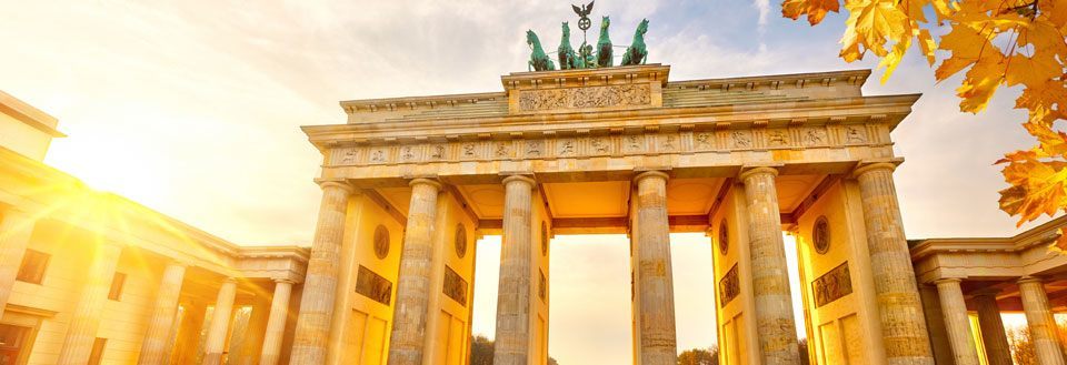 Bilden visar Brandenburger Tor i Berlin i det gyllene ljuset från solnedgången med höstlöv i förgrunden.