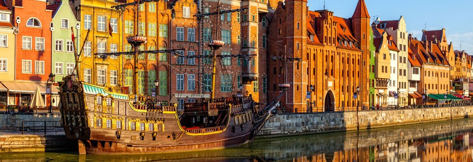 Färgglada byggnader speglar sig i vattnet i Gdansk bredvid ett gammalt segelfartyg i hamnen.