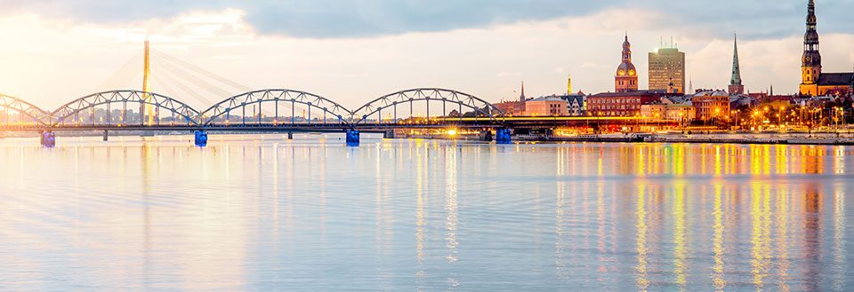 Bild av Riga vid floden i skymningen med ljus som reflekteras i vattnet och en bro i bakgrunden.