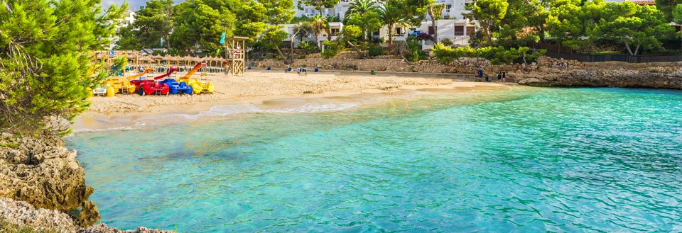 Bilden visar en pittoresk strand med turkosblått vatten, färgglada solstolar och grönskande träd vid kusten.