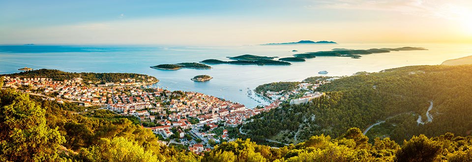 Panoramavy över en pittoresk kuststad i solnedgången, omgiven av gröna kullar och öar i fjärran.