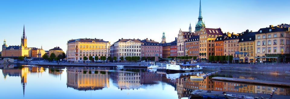 Panoramavy över en stad vid vattnet i gryningen med byggnaders spegling i vattnet.