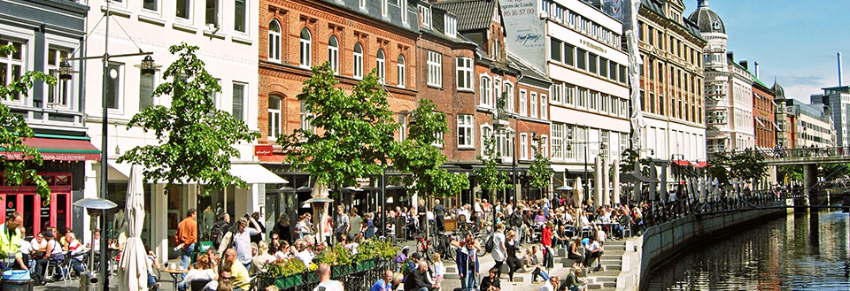 Glade mennesker nyder livet ved åen i Aarhus, bro och gröna träd under solig himmel.