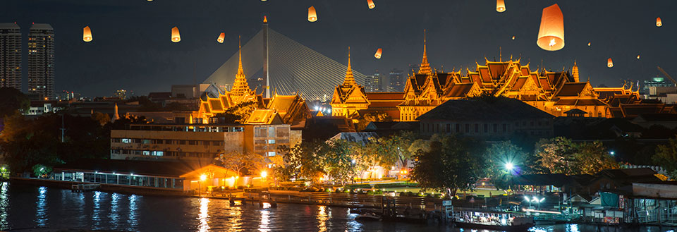 Bilden visar en flod om natten med traditionella lyktor som svävar i luften över ett thailändskt tempel.