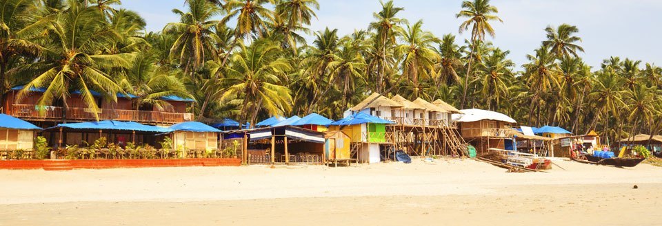 En tropisk strand i Goa med kokospalmer i bakgrunden och små stugor framför. Några båtar ligger på sanden.