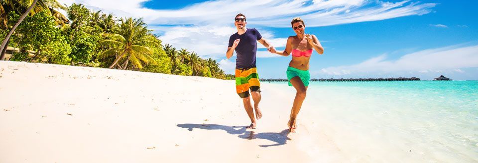 Två personer springer leende på solig strand med vit sand och palmer i bakgrunden.