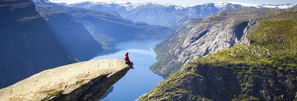 En person sitter ensam på kanten av stor klippavsats med en hänförande utsikt över fjorden och bergen.