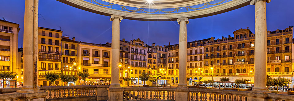 En stämningsfull kvällsbild av ett torg i Pamplona omgivet av klassiska byggnader som lyses upp av gatubelysning.