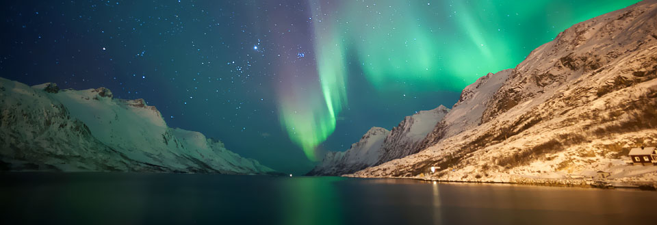 Norrsken lysar upp snötäckta berg och en stilla sjö med ett litet hus vid stranden.