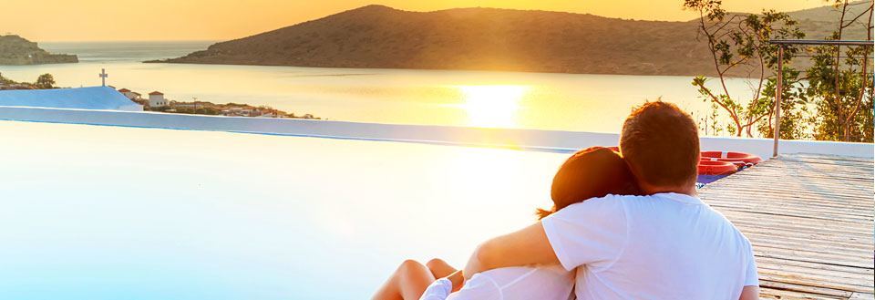 Ett par i avslappnad pose vid en infinitypool med utsikt över solnedgången och havet.
