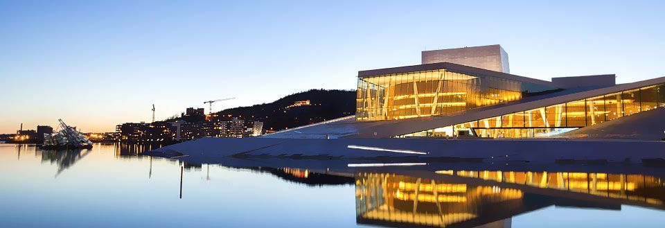 Modernt byggnadsverk speglar sig i lugnt vatten vid skymning med stadsljus i bakgrunden.